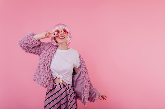 Cheerful girl in shiny peruke posing with sincere smile on pink background Portrait of pretty female model in fur jacket and stylish pants looking up