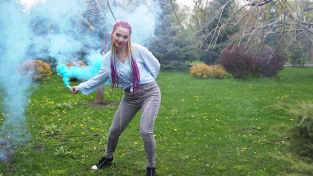 A cheerful girl in a shirt and jeans with bright rainbow braids and unusual makeup. She dances hiding in thick blue artificial smoke against the backdrop of spring park