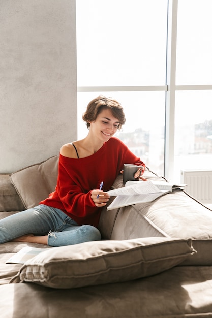 Cheerful girl studying at home while laying on a couch and drinking tea