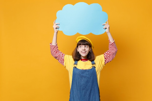 Cheerful girl teenager in french beret, denim sundress hold blue empty blank Say cloud, speech bubble isolated on yellow wall background. People sincere emotions lifestyle concept. Mock up copy space.