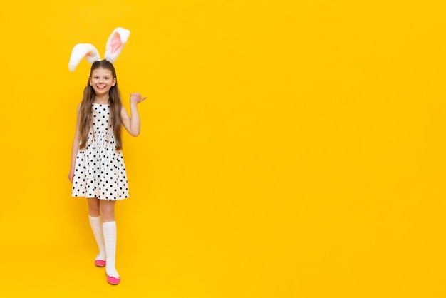 A cheerful girl with rabbit ears on her head and chicken eggs in her hands on a yellow background pointing to the side of the advertisement a funny happy child Easter child Easter holiday