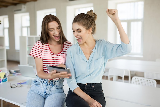 Cheerful girls sitting on desk happily screaming while using tablet together with modern empty office on background