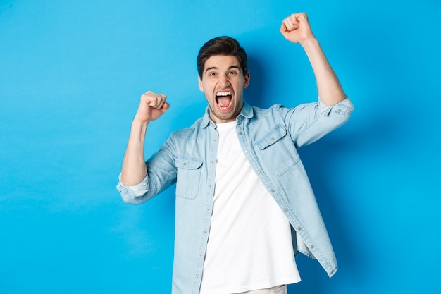 Cheerful guy making fist pumps and rooting for someone, shouting for joy, triumphing over win, standing against blue wall