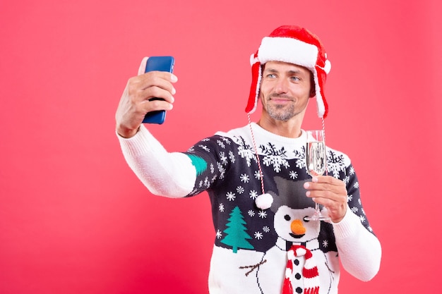 Cheerful guy making selfie on smartphone on red background. express positive emotions. drinking champagne. cheers. merry christmas. celebrate new year holiday. man in winter sweater and red hat.