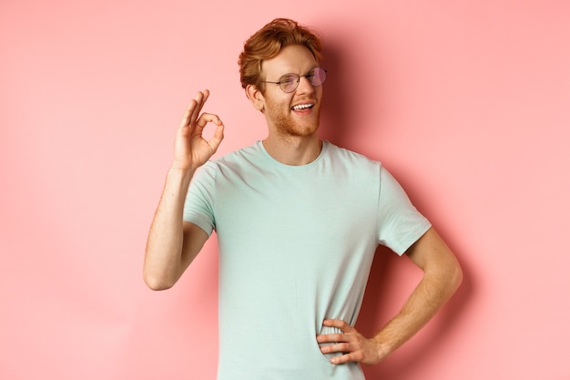 Cheerful guy with red hair and beard, wearing glasses, showing OK sign in approval and winking, smiling satisfied, standing over pink background