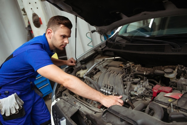 Cheerful, handsome, and confident car repair specialist in overalls repairs and replaces old parts with new ones in a car on a lift in service