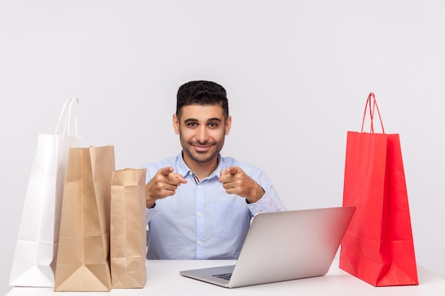 Cheerful handsome man sitting at desk with laptop surrounded by packages pointing to camera smiling recommending internet shopping purchase in online store studio shot isolated white background