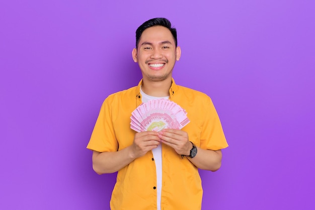 Cheerful handsome young Asian man holding a fan of money banknotes isolated on purple background