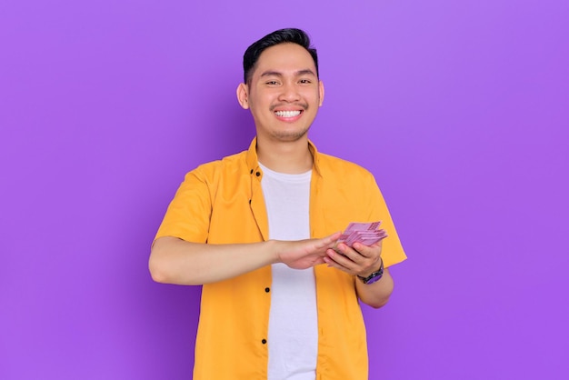 Cheerful handsome young Asian man throwing money banknotes isolated on purple background