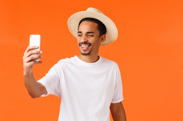 Cheerful happy african-american guy taking selfie on vacation under sun near sandy beautiful beach tropical island, holding phone and smiling at mobile , standing orange wall