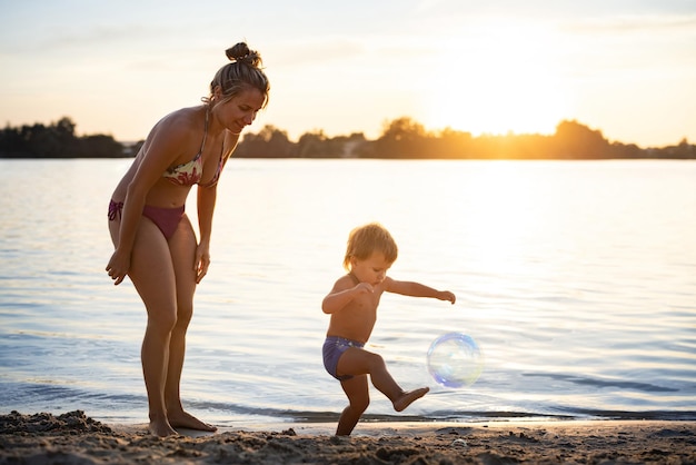 Cheerful happy beautiful mother in modern bikini swimsuit plays with her little funny baby inflating bright multicolored soap bubbles for him, while enjoying sunny summer holidays