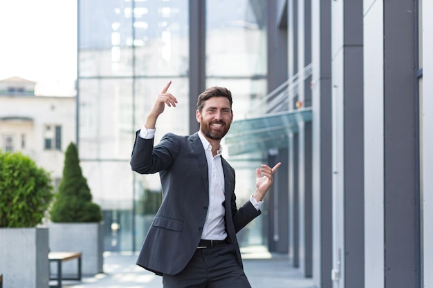 Cheerful happy business man dancing walk the city street background a modern office building outside, outdoors Funny successful businessman in suit celebtes victory