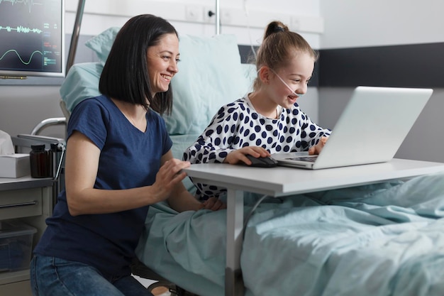 Cheerful happy mother enjoying time with smiling daughter while in clinic ward room. Relaxed mother and daughter sitting in pediatric clinic patient ward room while using modern computer.