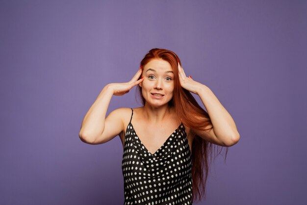 A cheerful happy woman rejoices in winning, laughs and is pleasantly shocked. Emotional red-haired girl in a black polka-dot dress on a lilac lavender wall