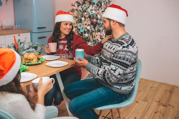 Cheerful and happy young woman sits at table with husband nd daughter. She looks at him and touching his shoulder. Guy looks at her. They hold cups in hands. People wear red hats.