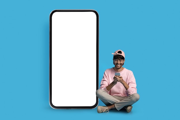 Cheerful hindu guy hipster sitting by huge smartphone