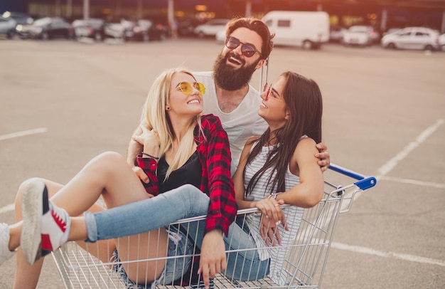 Cheerful hipsters having fun with shopping trolley on parking lot