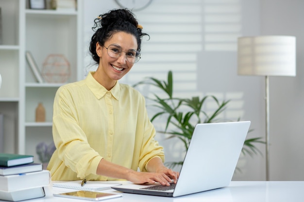 A cheerful hispanic woman wearing glasses works on a laptop in a welllit room surrounded by books