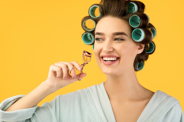 Photo cheerful housewife curling eyelashes and hair posing over yellow background