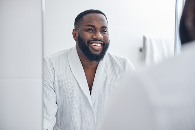 Cheerful international brunette man keeping smile on his face while looking at his reflection in glass