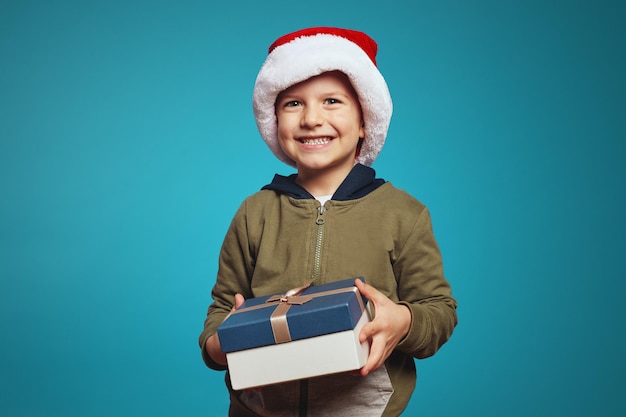 Cheerful kid boy in christmas hat opening a gift box isolated over blue