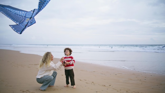 Cheerful kid playing kite with mother on beach caring young parent helping son