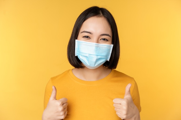 Cheerful korean woman in medical face mask support people during pandemic wear personal protective equipment from covid19 showing thumbs up in approval yellow background