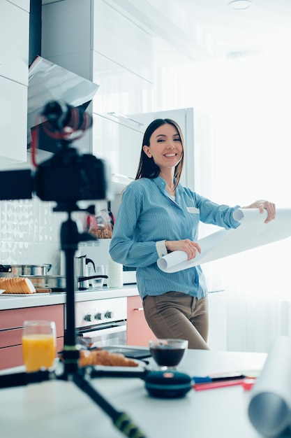 Cheerful lady holding a paper roll in the kitchen and smiling to the camera on tripod