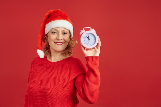 Cheerful lady in santa hat holds clock isolate on red background. Merry Christmas.