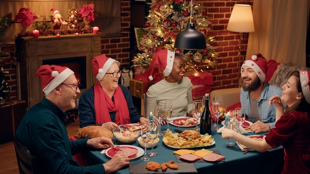 Cheerful laughing heartily multiethnic family toasting sparkling wine while singing Christmas songs at dinner. Festive diverse people wearing traditional hats enjoying winter holiday together at home.