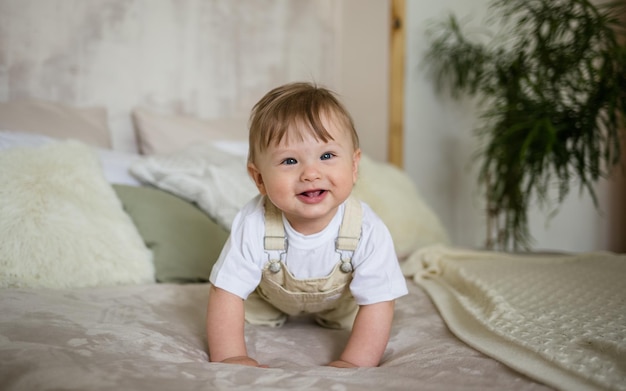 A cheerful little boy in a beige jumpsuit crawls on the bed in the room Learn to crawl