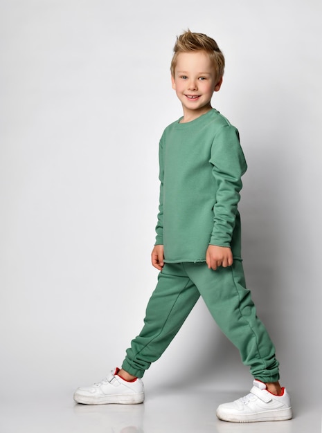 Cheerful little boy in a comfortable sports suit of green color stands halfsided and joyfully looks at the camera against a light background