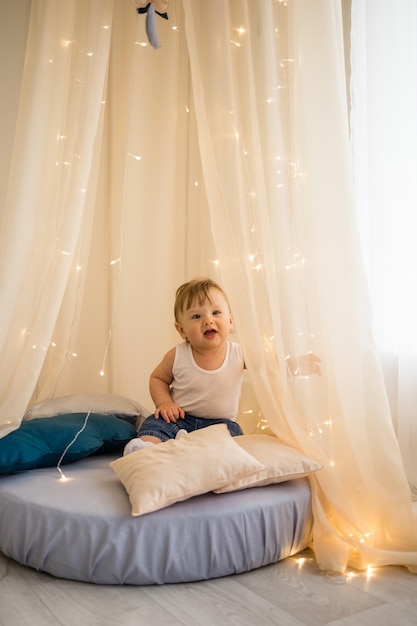 A cheerful little boy is sitting on a mattress under a canopy and playing Caucasian baby boy sitting in the nursery