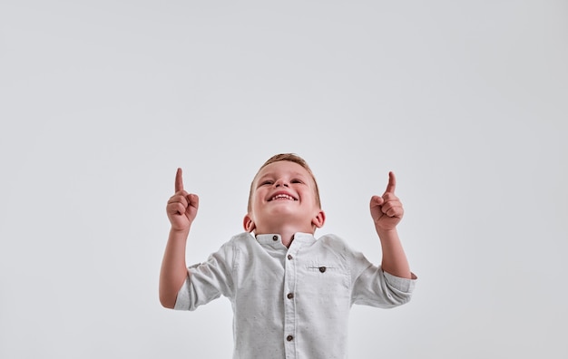 Cheerful little boy looking and pointing up with both hands over gray background and smiling.