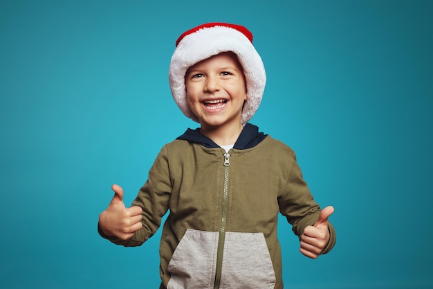 Cheerful little caucasian boy wearing christmas hat showing thumbs up over