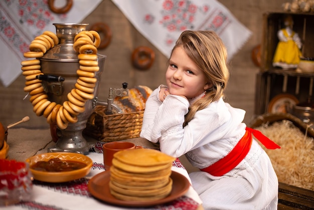 Cheerful little girl in folk costume seating at the table with samovar while celebrating Maslenitsa