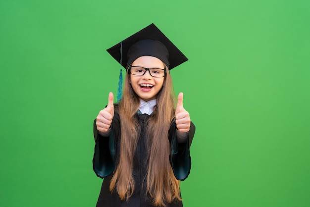 A cheerful little girl in a graduation ceremonial robe and a cap shows a gesture with her fingers everything is fine on a green isolated background.