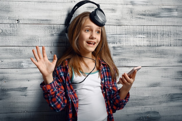 Cheerful little girl in headphones listen to music in studio. Children and gadget, kid isolated on wooden background, child emotion, schoolgirl photo session