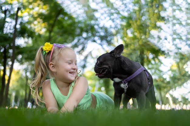 Cheerful little girl lies on the lawn in the park and looks at the french bulldog who is next to her