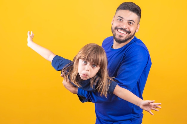 Cheerful little girl playing with her father on yellow background stretching hands pretending to fly Young father holding his daughter having fun together