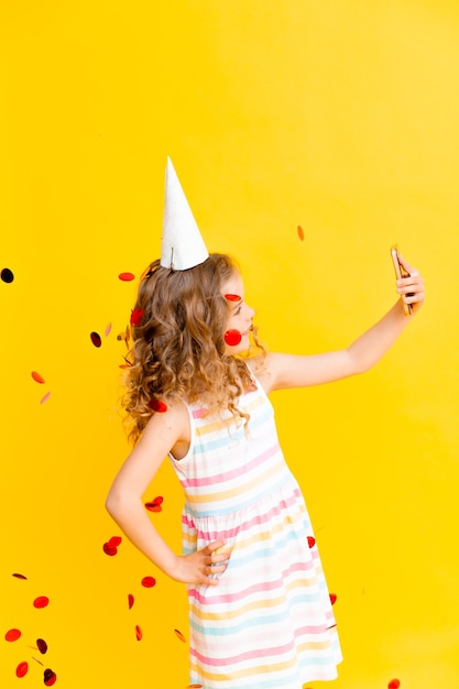 Cheerful little girl with blond curly hair is celebrating her birthday. The child holds the phone, takes a selfie in the rain of confetti. Close-up portrait on yellow background.