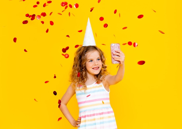 Cheerful little girl with blond curly hair is celebrating her birthday. The child holds the phone, takes a selfie in the rain of confetti. Close-up portrait on yellow background.