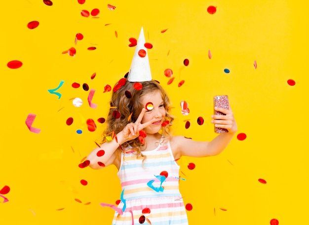 Cheerful little girl with blond curly hair is celebrating her birthday. The child holds the phone, takes a selfie in the rain of confetti. Close-up portrait on yellow background.