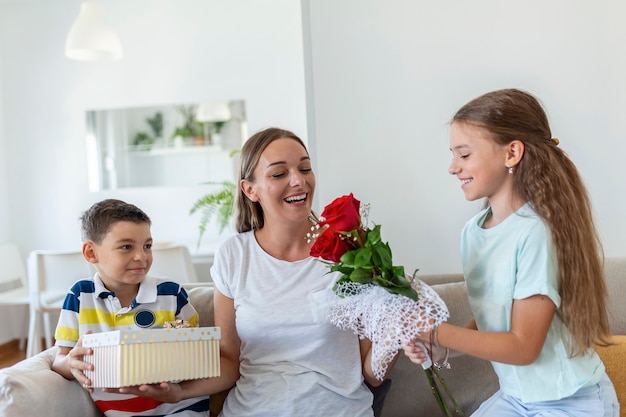 Cheerful little girl with bouquet of roses flowers and youngest brother with gift box smiling and congratulating happy mom on mother day at home. Happy Mothers Day!