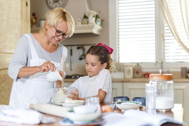 Cheerful little girl with her grandmother cooking together at kitchen table. Horizontal view.