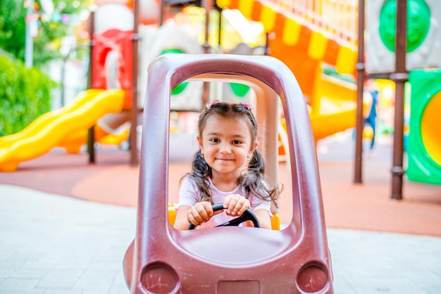 Cheerful little girl with two ponytails sits in a childrens car on the playground in the park