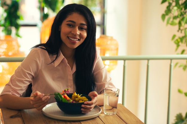 Cheerful long haired young woman looking glad and smiling while sitting with a bowl of vegetable meal and a glass of water