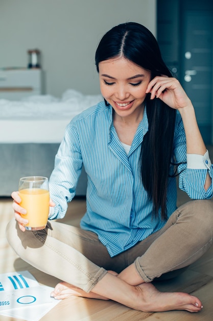 Cheerful long haired young woman sitting on the floor with a glass of orange juice and smiling Graphic charts by her side