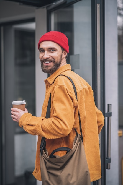 Cheerful look. Man in a red hat and orange jacket with a coffee cup in hands looking cheerful