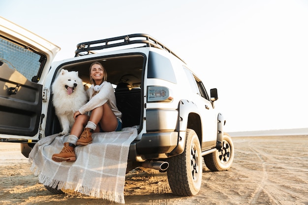 Cheerful lovely girl playing with her dog while sitting in a car at the beach